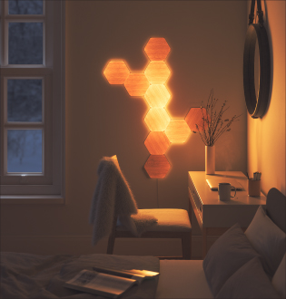 This is an image of a 10 panel Nanoleaf Elements layout mounted onto the wall beside a desk in a bedroom. Modular smart light panels that are secured to the walls with included adhesives and connected together with linkers. The wood look hexagons are the perfect decorative piece that adds style and personality in the bedroom while doubling as a customizable ambient light.
