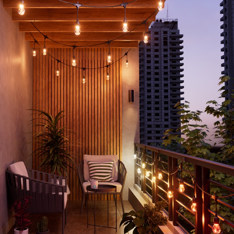 Outdoor string lights emitting a warm white glow on an apartment patio with a view of skyscrapers and trees. Wooden frame decal on the wall with two chairs, a small black circular table, and a few plants. Wooden frames on the patio ceiling supporting the lights.